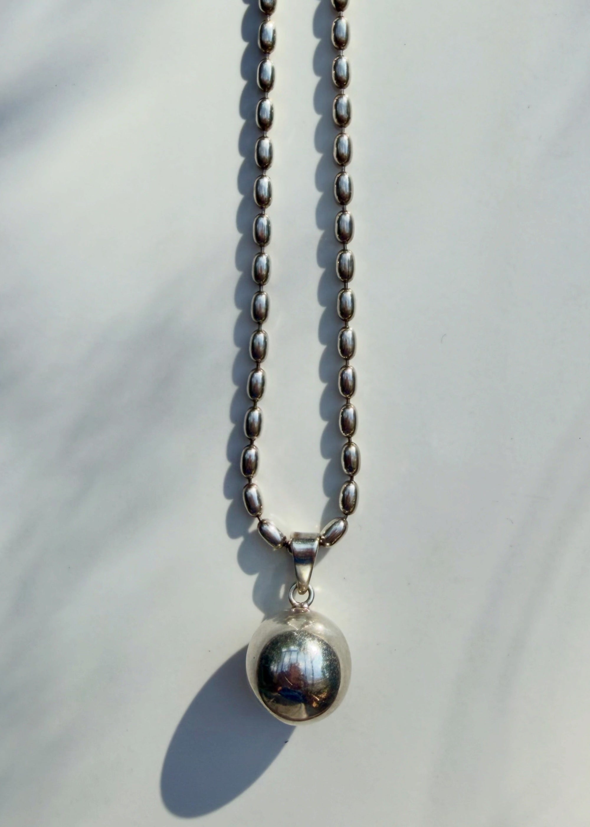 A silver beaded chain necklace with a polished spherical pendant, photographed against a light gray background with soft leaf shadows.