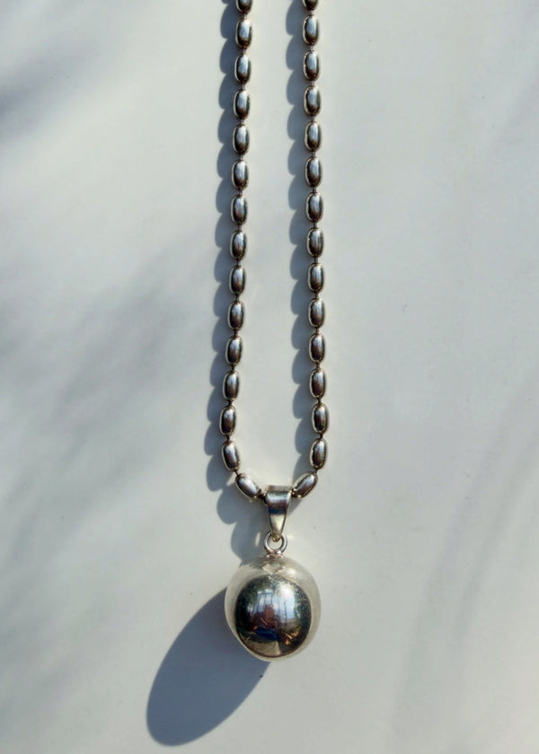 A silver beaded chain necklace with a polished spherical pendant, photographed against a light gray background with soft leaf shadows.