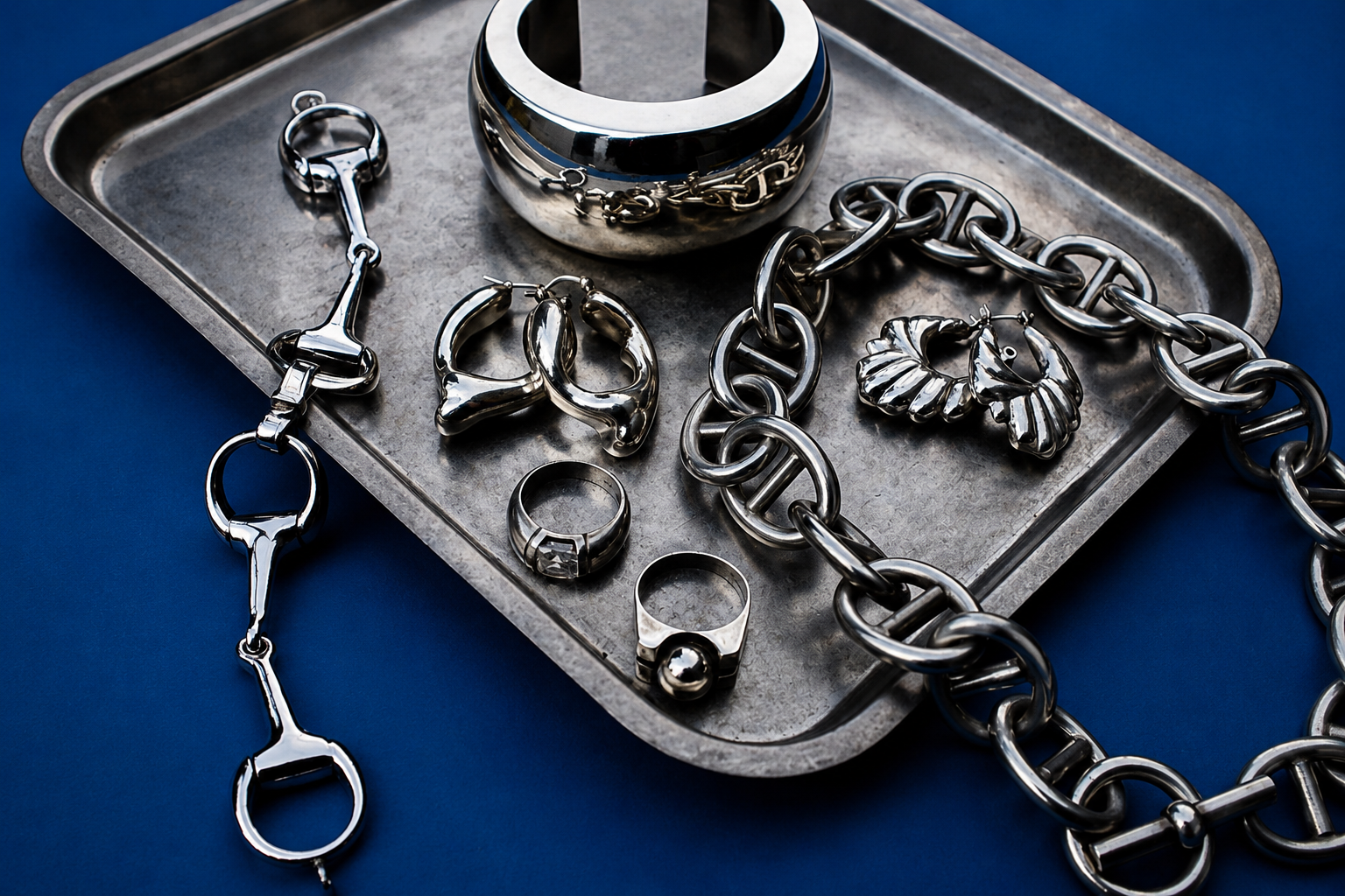 Collection of silver jewelry including rings and a chain on a metal tray with a blue background.
