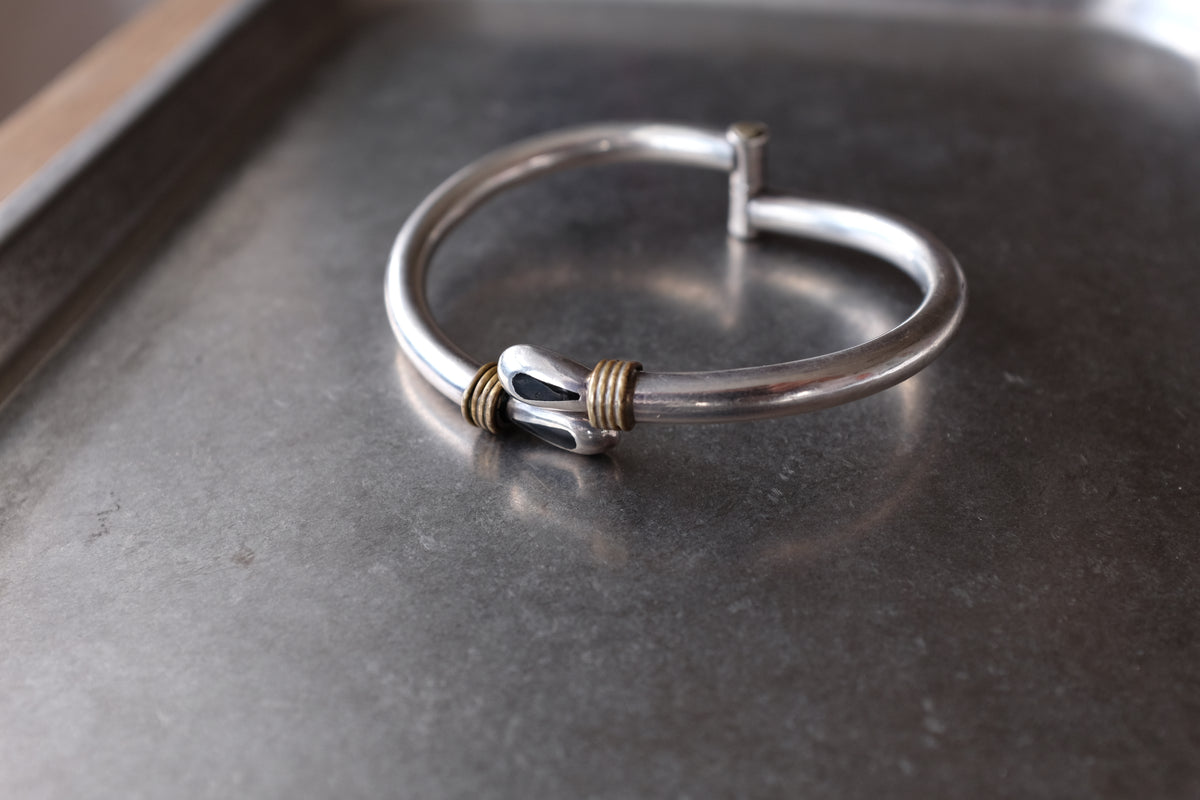 A sterling silver open cuff bracelet with a decorative toggle clasp and two small gold wire-wrapped accents, resting on a dark gray metal surface.