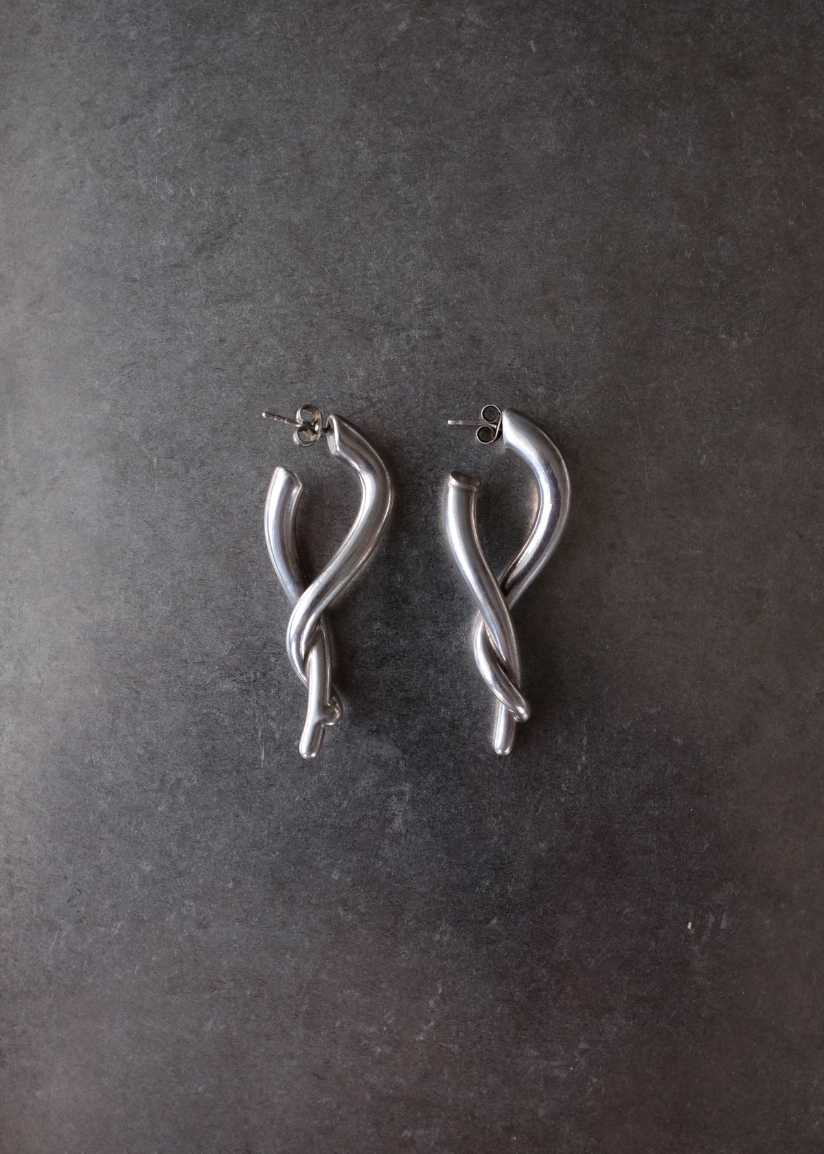 A pair of silver-toned twisted hoop earrings with post backings, displayed on a dark textured slate surface.