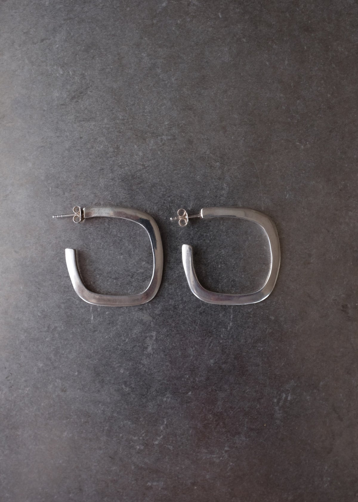 A pair of silver-toned square hoop earrings with post backs, displayed on a dark textured gray surface.