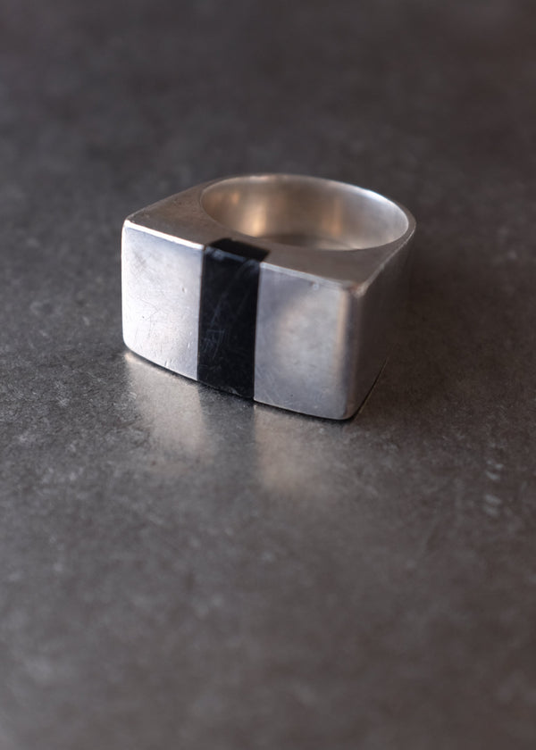 A silver-toned geometric ring with a central black stripe, resting on a dark textured surface.
