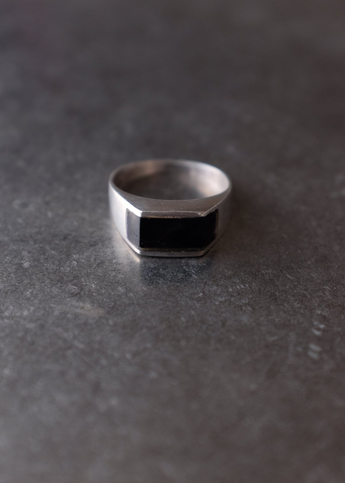 A silver-toned metal ring with a black rectangular inlay, photographed on a dark textured gray surface.