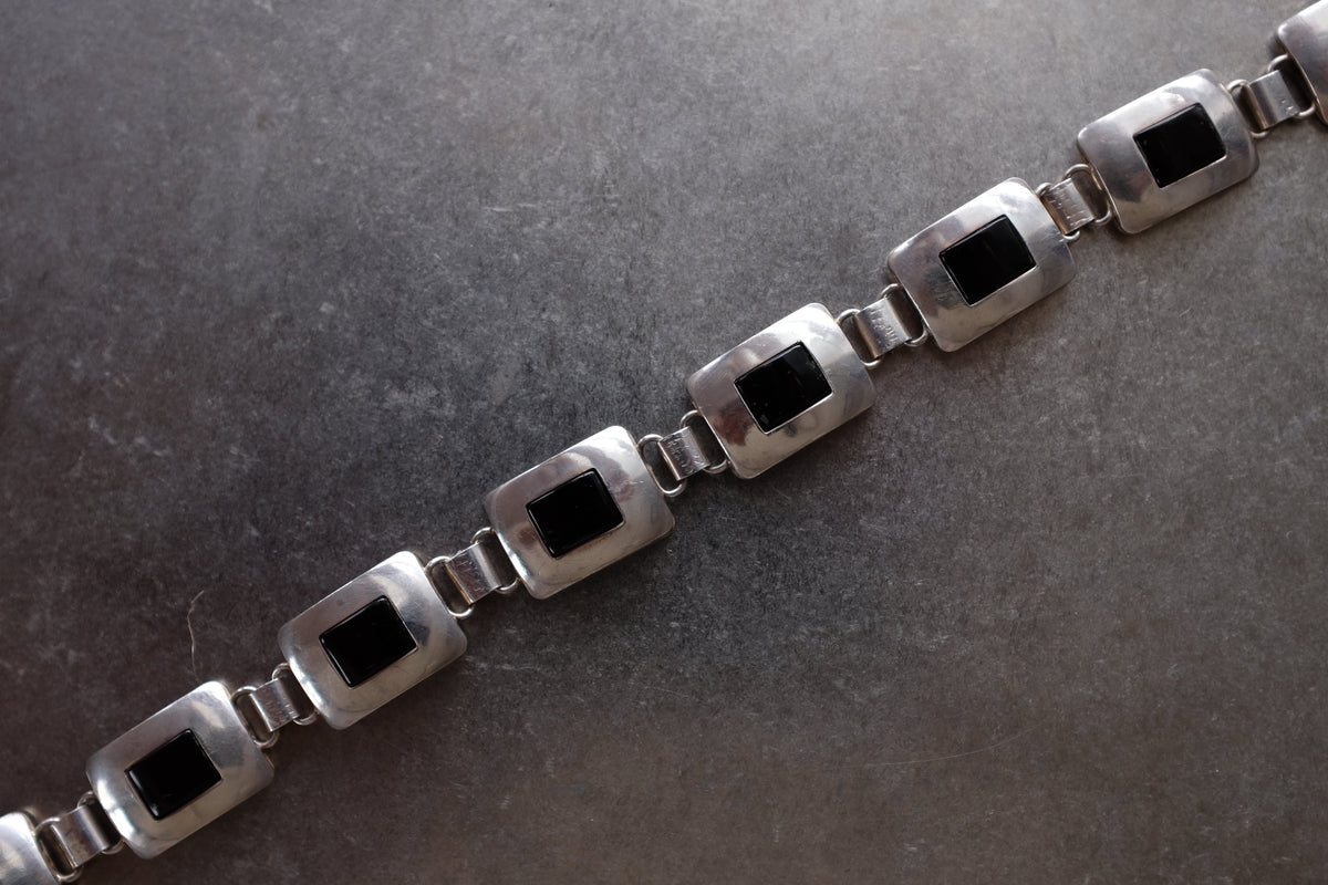 A close-up of a silver-toned chain bracelet with rectangular links featuring dark inlay centers, laid diagonally on a dark textured surface.