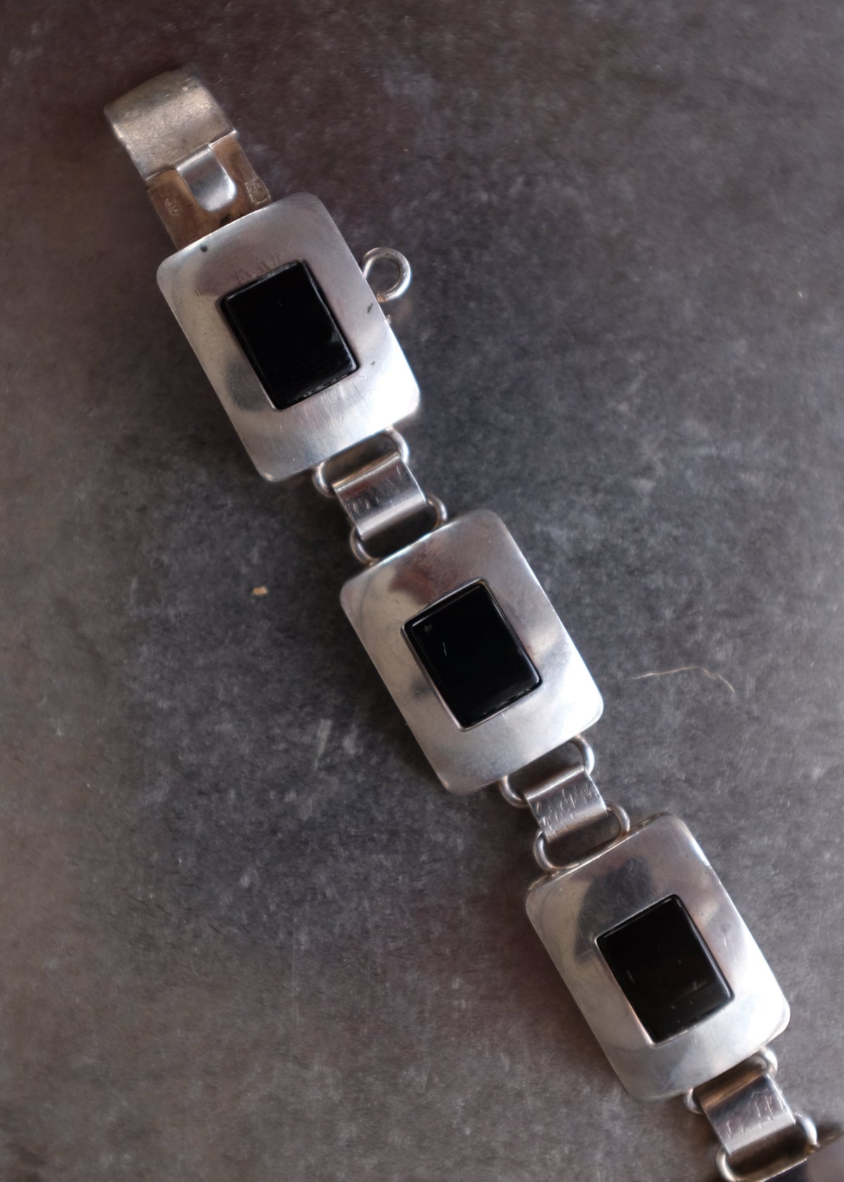 A silver-toned metal bracelet with three rectangular links, each featuring a square black inlay, photographed on a dark textured surface.