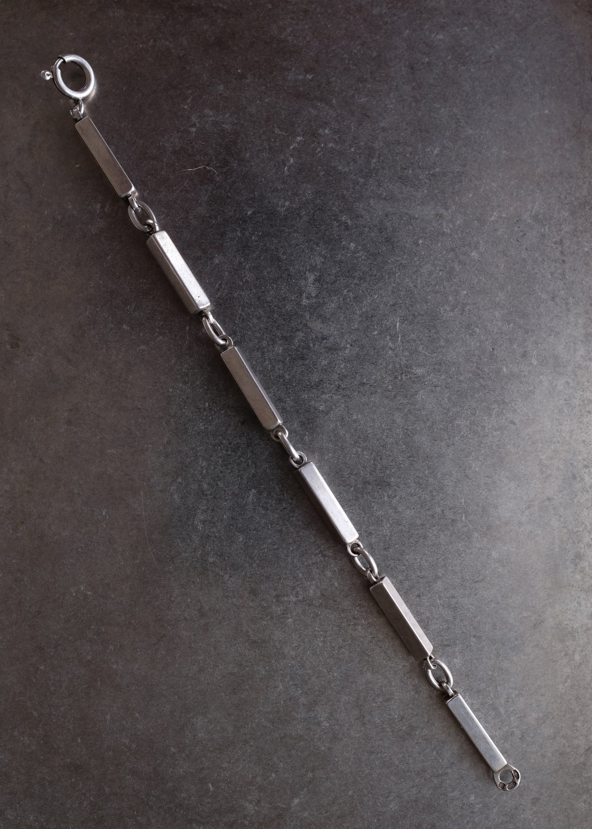 A silver-toned metal link chain bracelet with elongated rectangular links and oval ring connectors, laid diagonally on a dark textured surface.