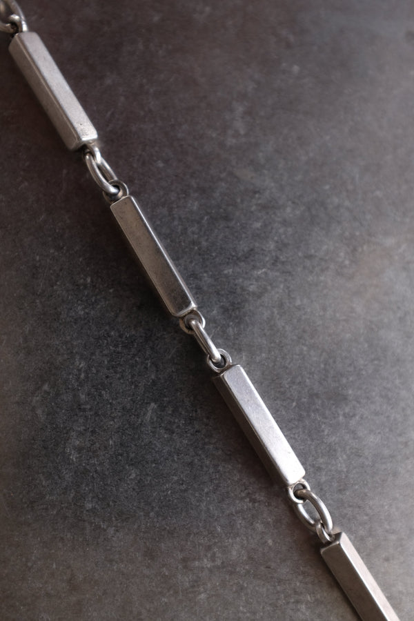 A close-up of a silver-toned metal chain with rectangular links and small oval connectors, laid diagonally on a dark textured surface.
