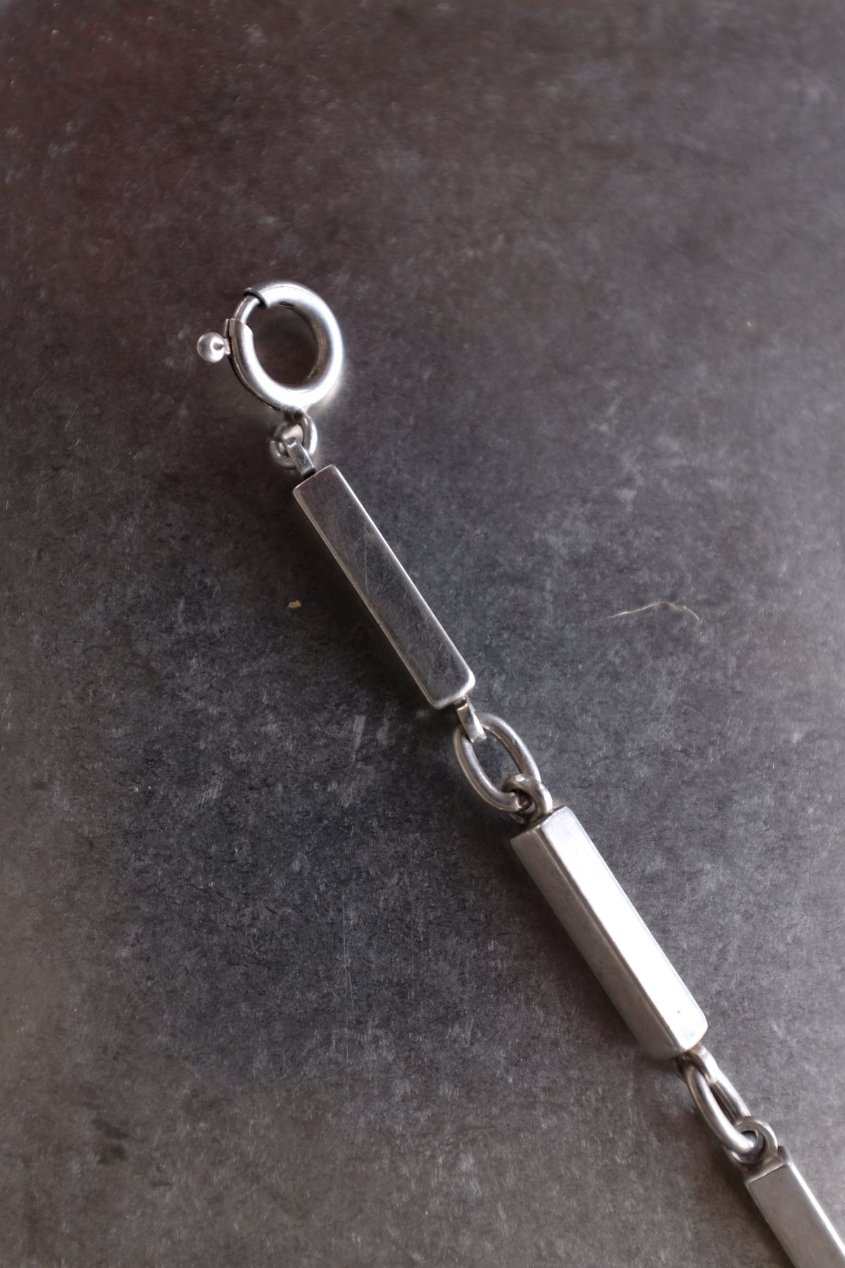 A close-up of a silver-toned metal chain link bracelet showing polished rectangular links, a connector ring, and a lobster clasp on a dark textured background.