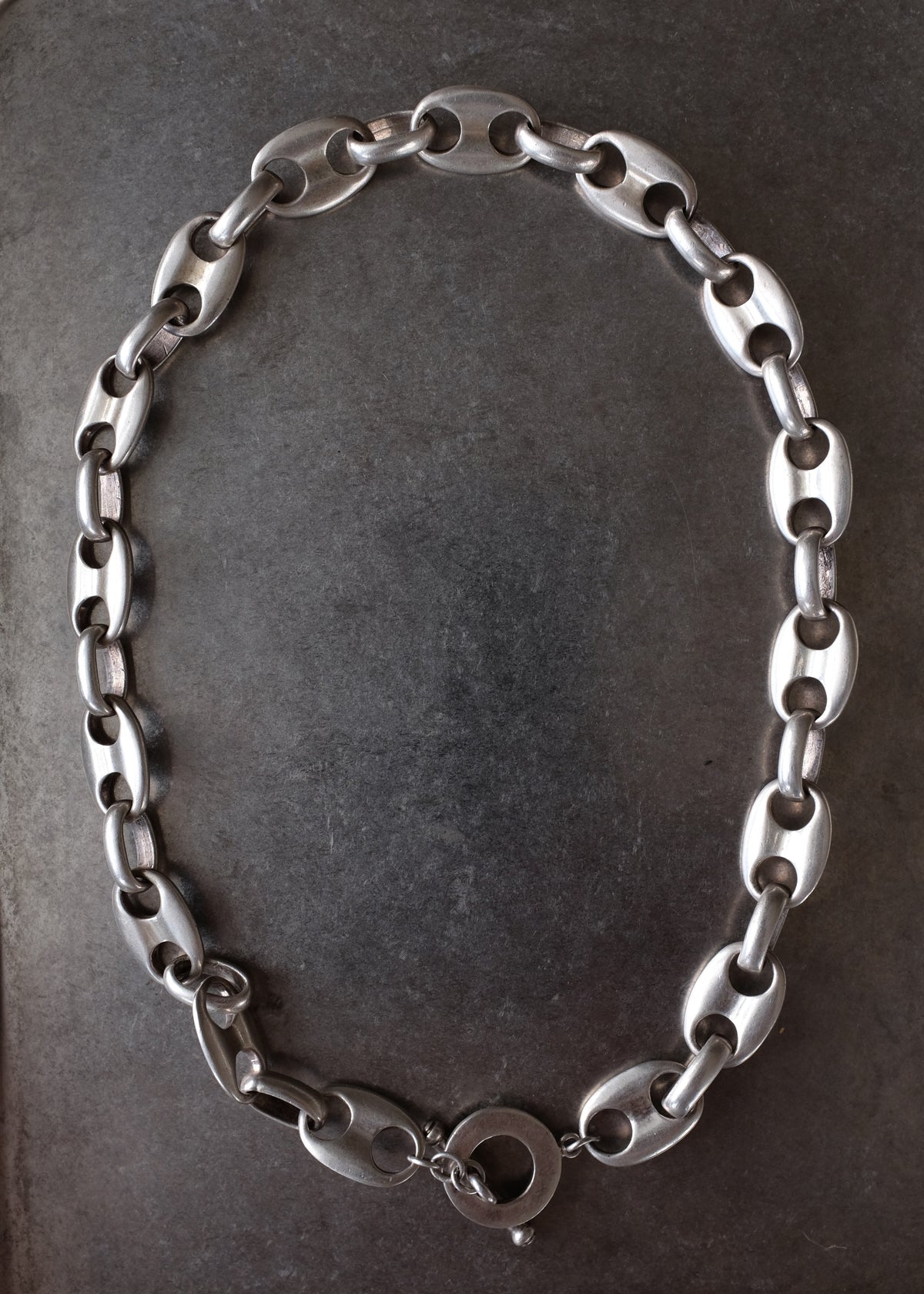 A silver-toned metal chain necklace with elongated oval links and a circular clasp, arranged in an oval shape on a dark textured surface.