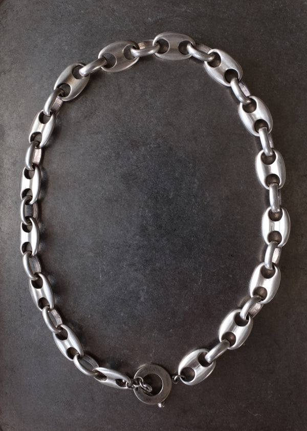 A silver-toned metal chain necklace with elongated oval links and a circular clasp, displayed on a dark gray rectangular tray.