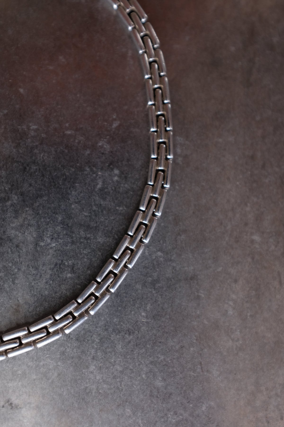 A close-up of a silver-toned metal link chain, likely a bracelet or necklace, arranged in a curve on a dark textured gray surface.