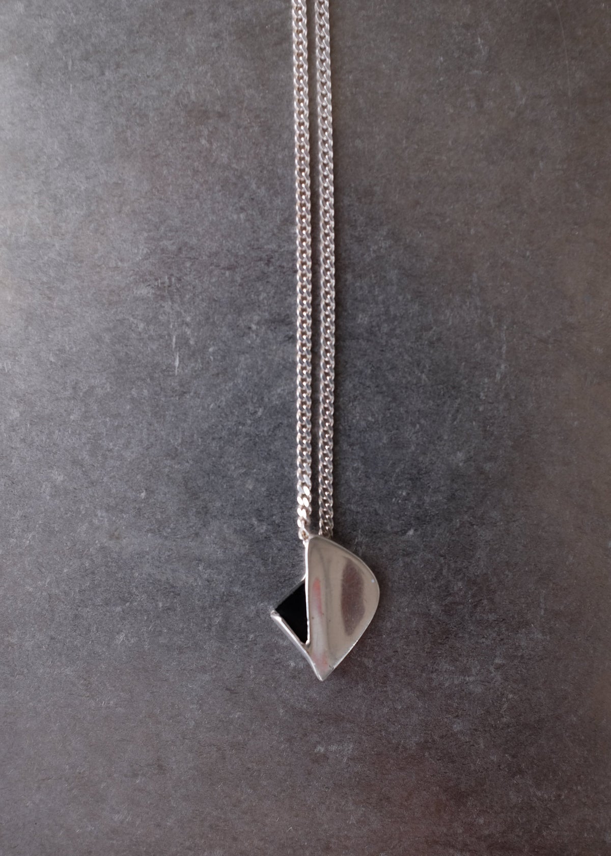 A close-up of a silver chain necklace with a polished teardrop-shaped pendant resting on a dark gray textured surface.