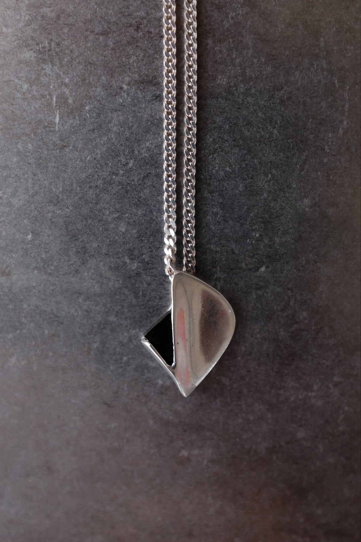 A silver pendant necklace with a polished, organic teardrop-shaped pendant and a small black triangular accent, hanging from a silver chain against a dark textured gray background.