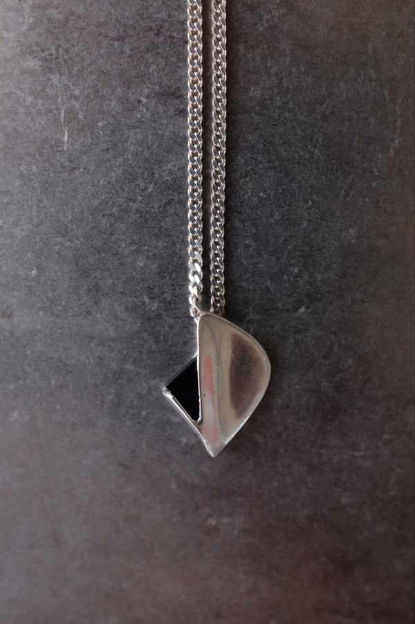 A silver pendant necklace with a polished, organic teardrop-shaped pendant and a small black triangular accent, hanging from a silver chain against a dark textured gray background.