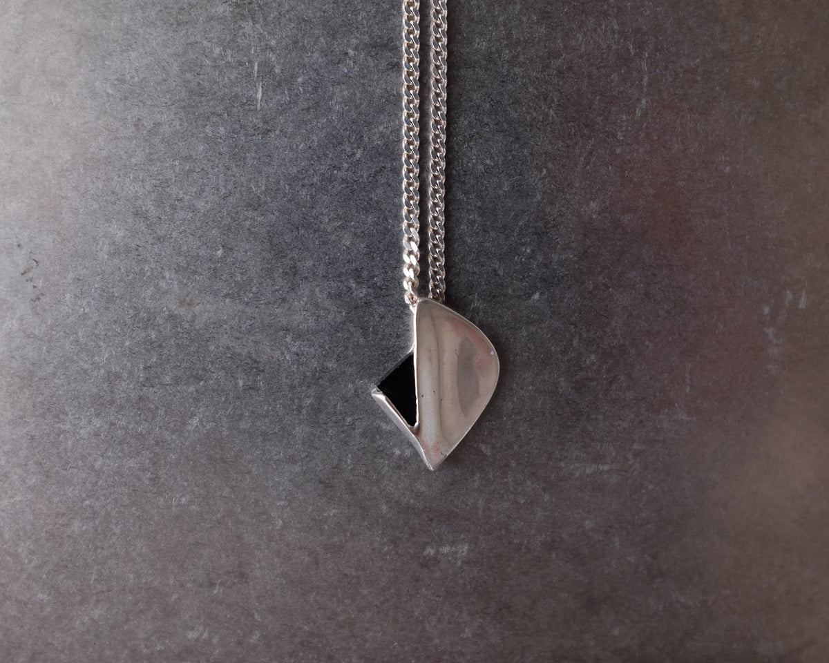 A silver geometric pendant necklace with a faceted triangular charm and a small black triangular accent, hanging from a silver chain against a textured dark gray background.