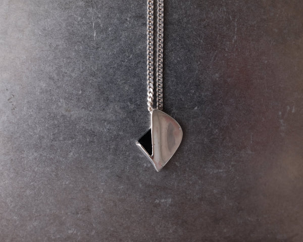 A silver geometric pendant necklace with a faceted triangular charm and a small black triangular accent, hanging from a silver chain against a textured dark gray background.