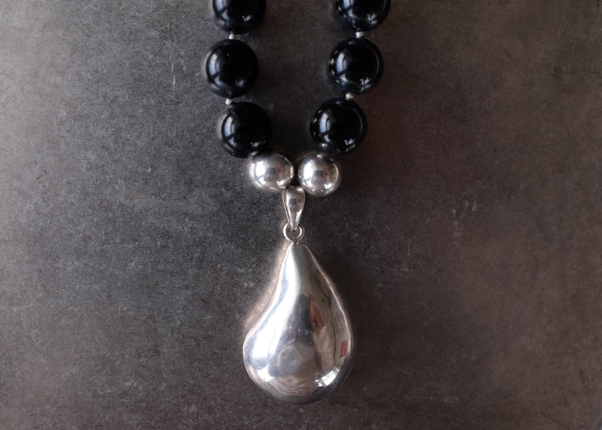 A close-up of a beaded necklace with glossy black round beads, small silver spacer beads, and a large polished silver teardrop pendant, resting on a dark gray surface.