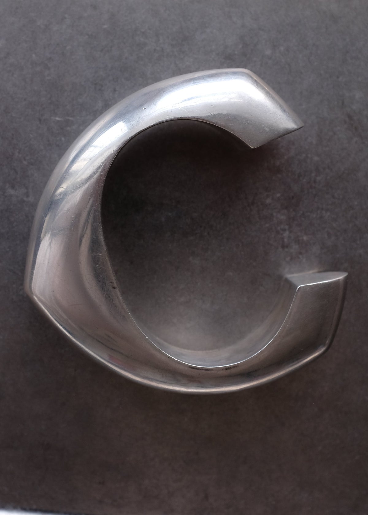 A polished silver metallic C-shaped ring or bangle with a smooth, reflective finish, shown on a dark gray textured background.