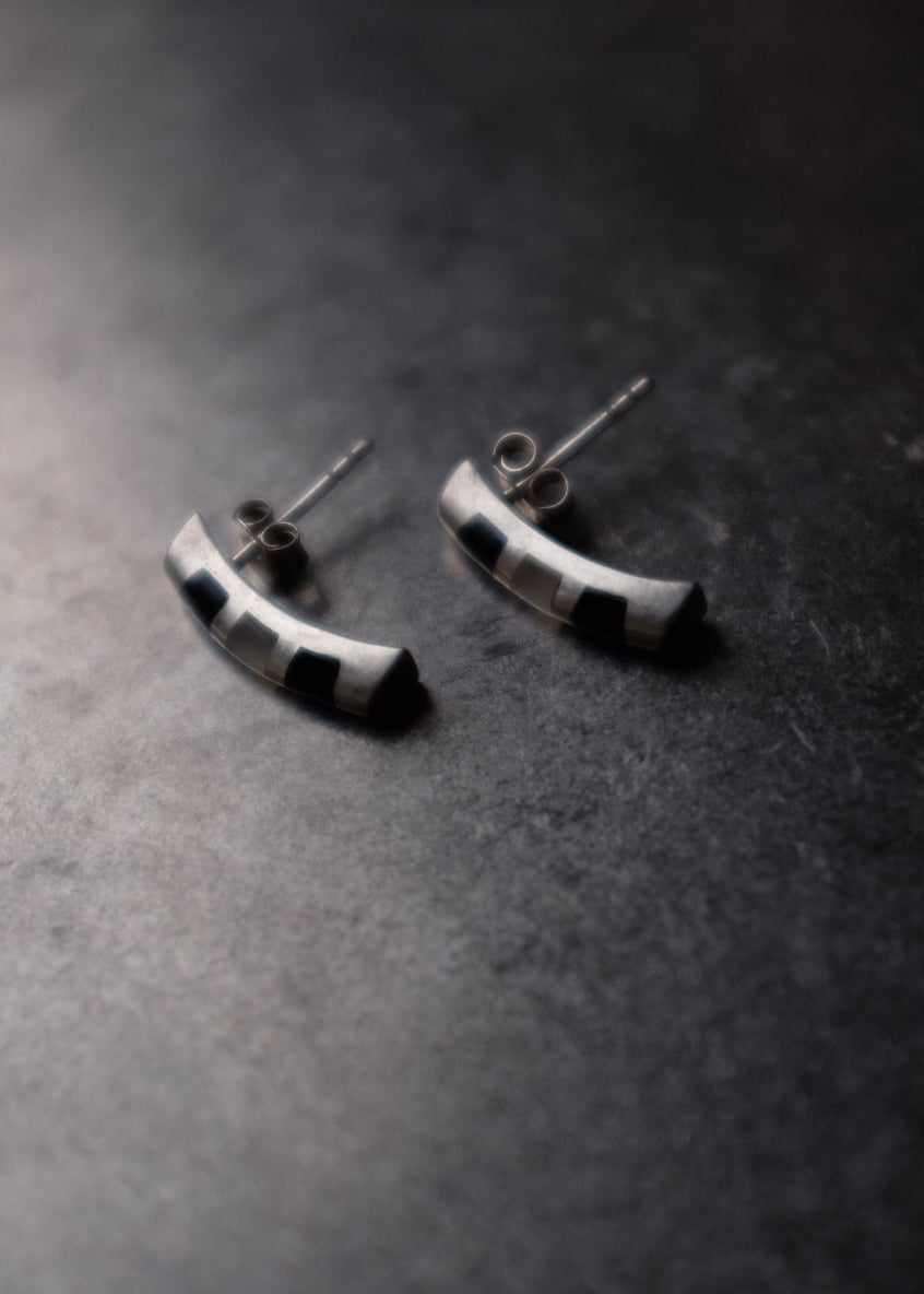 A pair of small silver crescent-shaped stud earrings with black inlay details, resting on a dark textured surface.