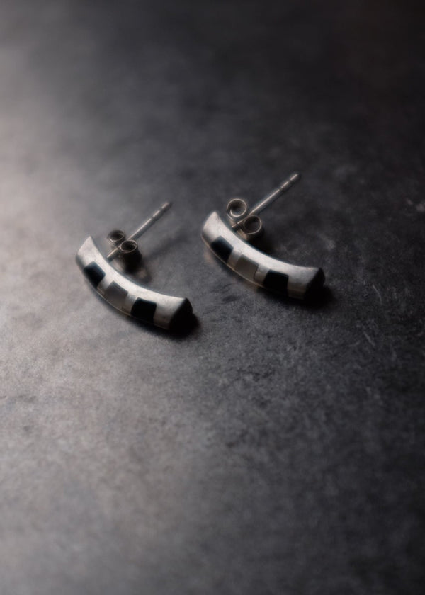 A pair of small silver crescent-shaped stud earrings with black inlay details, resting on a dark textured surface.