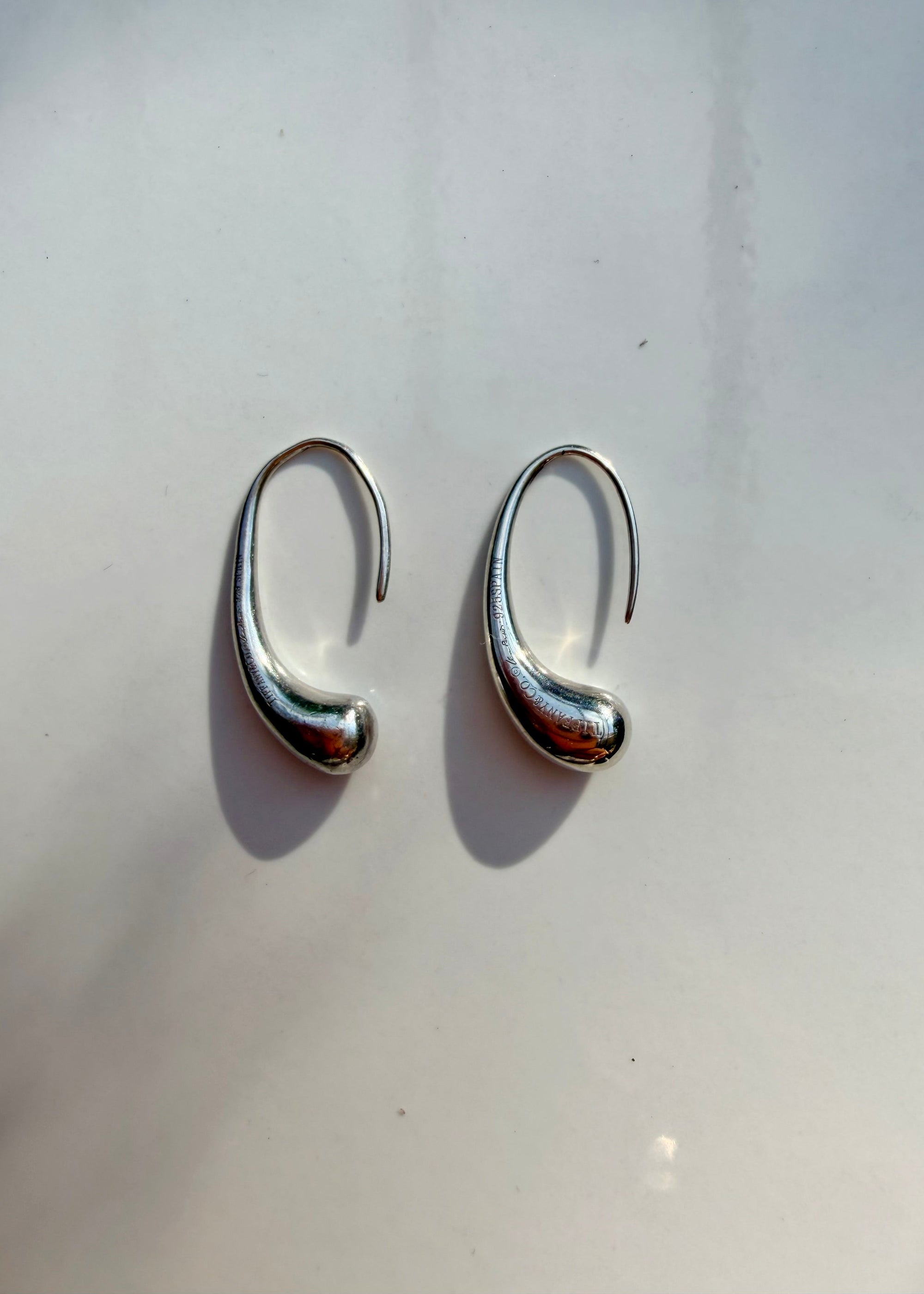 A pair of polished silver huggie hoop earrings with a curved, organic shape, displayed on a light off-white surface with soft shadows.
