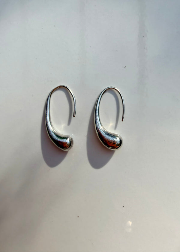 A pair of polished silver huggie hoop earrings with a curved, organic shape, displayed on a light off-white surface with soft shadows.