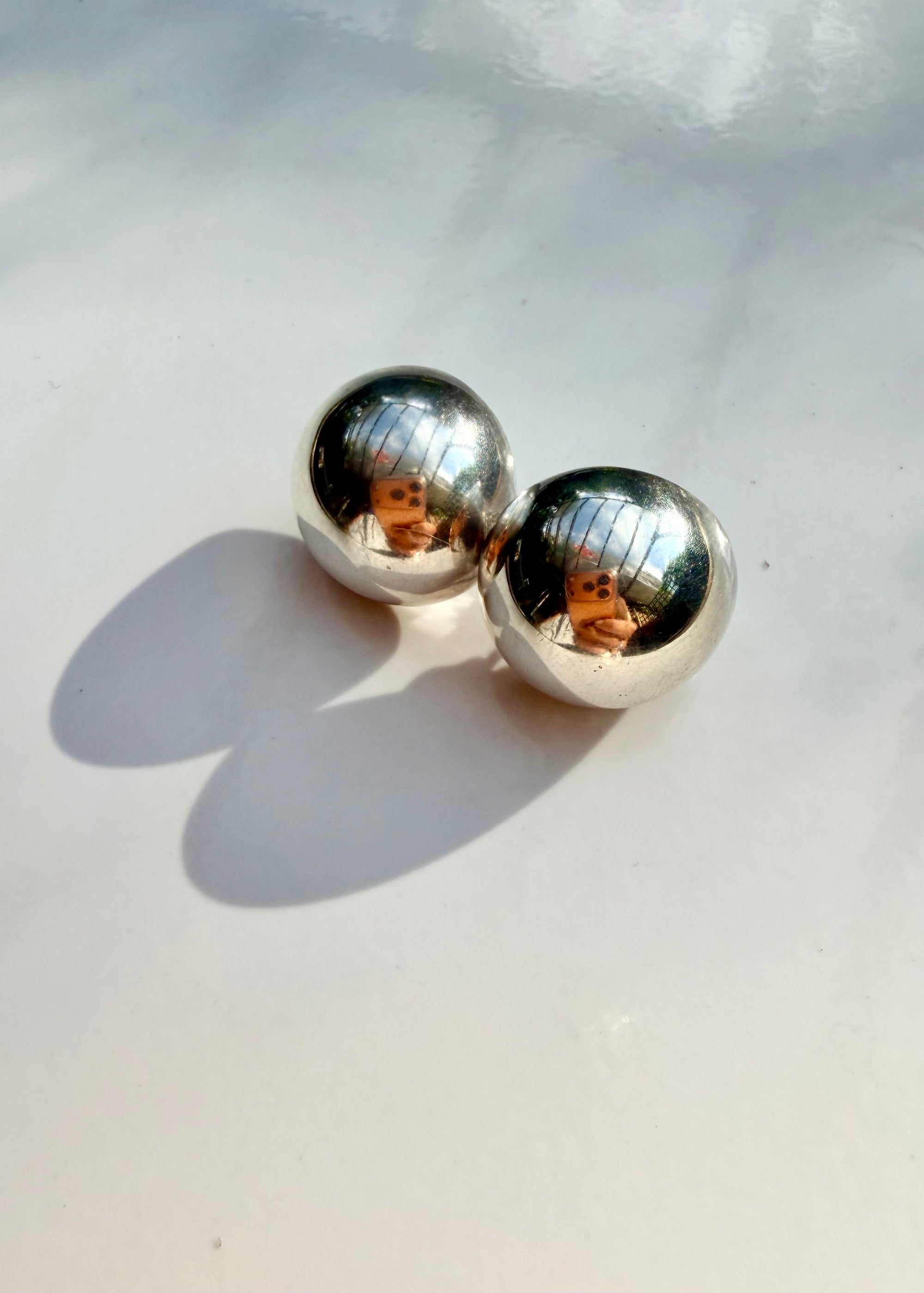 A pair of polished silver ball stud earrings resting on a light gray surface with soft shadows and reflections.