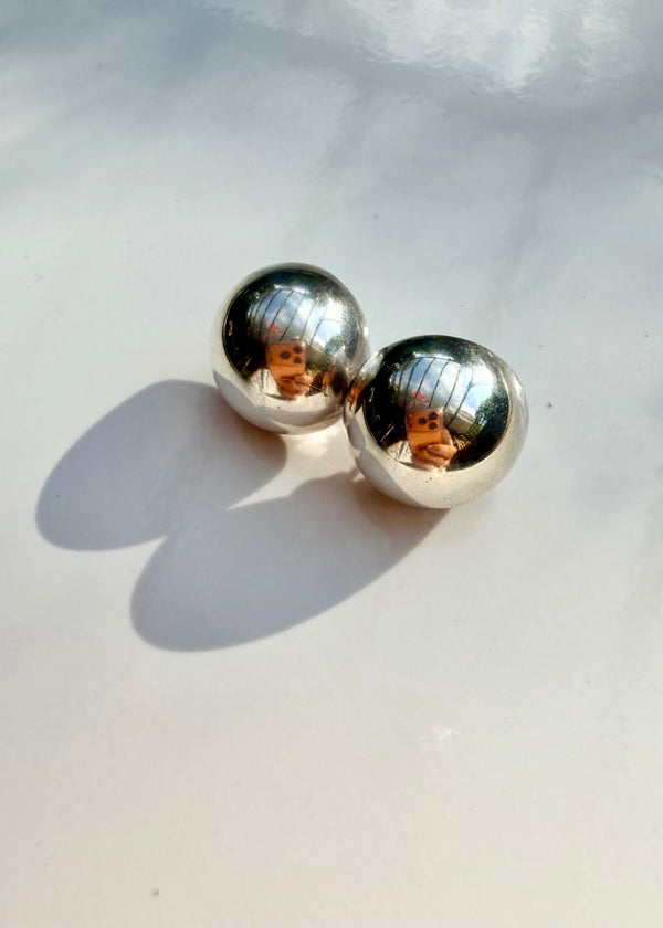 A pair of polished silver ball stud earrings resting on a light gray surface with soft shadows and reflections.