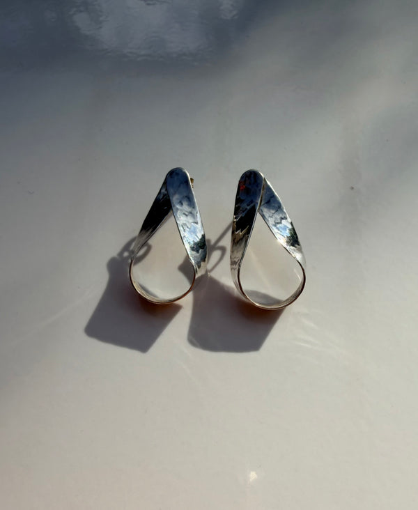 A pair of hammered silver teardrop-shaped hoop earrings resting on a light gray surface with soft shadows.