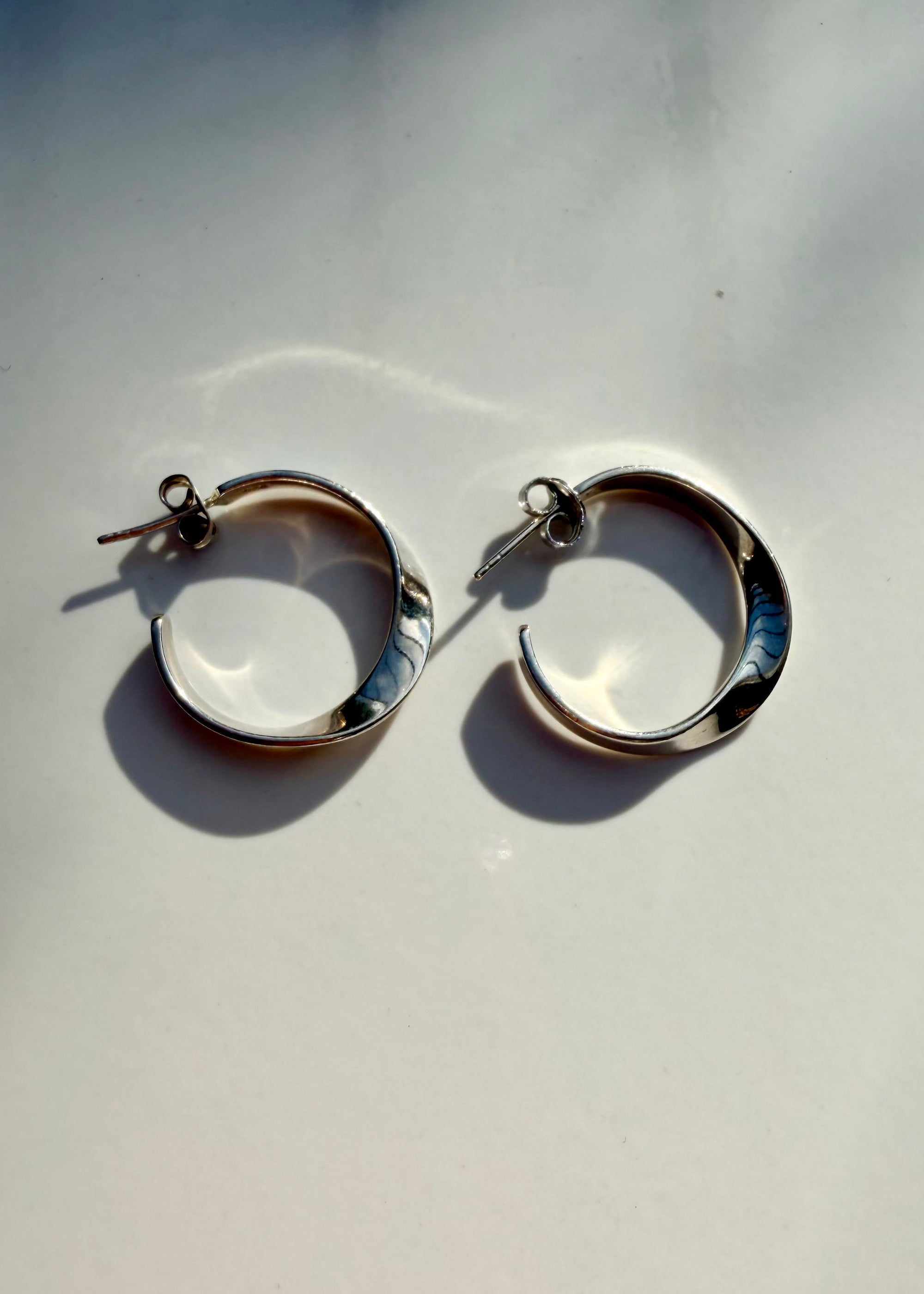 A pair of polished silver-tone hoop earrings with a twisted design, displayed on a light gray surface with soft shadows.