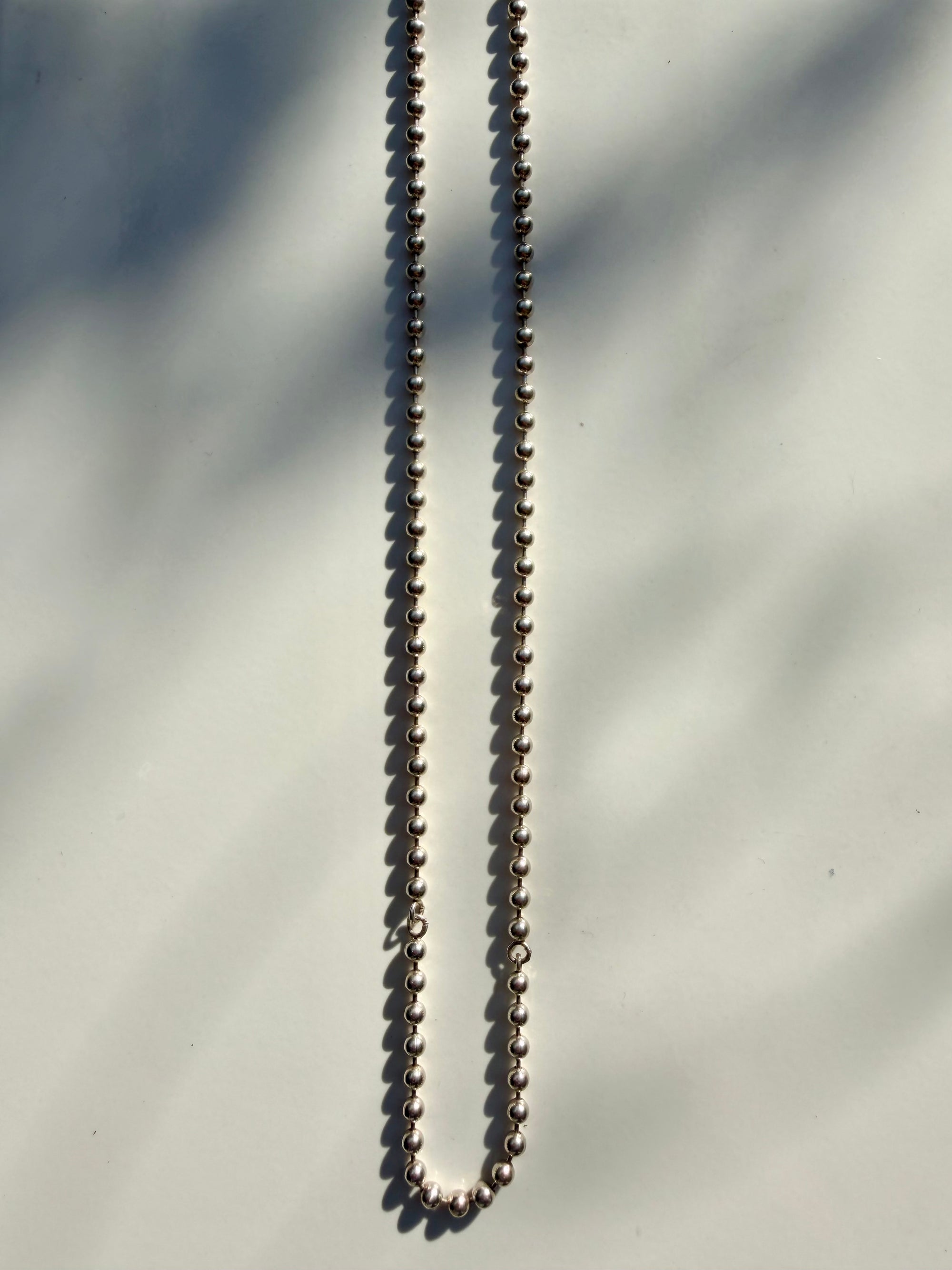 A silver ball chain necklace laid vertically on a light gray surface with soft leaf shadows cast across it.